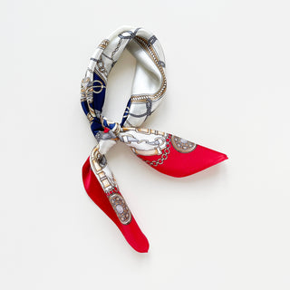 Red and navy silk bandana scarf with baroque equestrian pattern featuring ornamental chains, buckles, and floral designs, elegantly tied against white background.