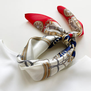Red & navy baroque silk bandana scarf with equestrian design featuring ornamental chains and buckles, displayed tied in a knot on white fabric.