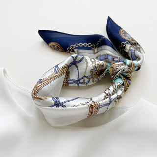 Luxury silk scarf in navy and white with ornamental equestrian design featuring decorative chains, buckles, and gold accents, elegantly folded on white surface.