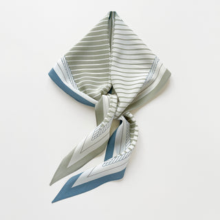 Diamond-shaped silk scarf featuring sage green stripes with blue accents and monogram detailing, elegantly knotted to showcase its luxurious crepe de chine texture