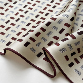 Ivory silk twill scarf with geometric block pattern in burgundy and blue, featuring elegant hand-rolled burgundy edges and luxurious draped texture.