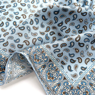 Light blue silk neckerchief with paisley pattern in blue and brown tones, featuring decorative border with intricate designs and hand-rolled edges.