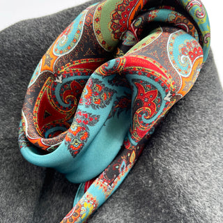 Folded turquoise silk scarf with intricate bohemian paisley pattern in vibrant red, orange, and green on gray background