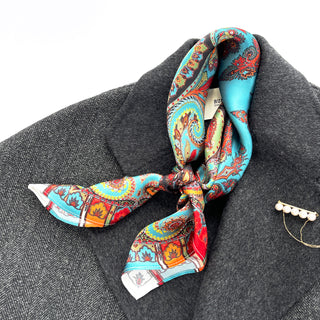 Turquoise and orange silk scarf with intricate paisley boho pattern, tied elegantly on a gray coat with pearl brooch accent