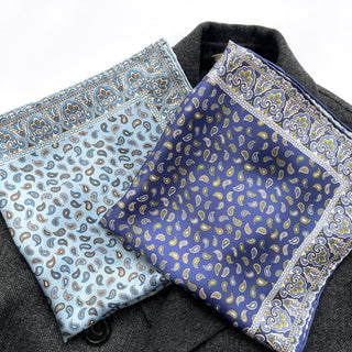 Two silk paisley neckerchiefs with hand-rolled edges - one sky blue and one rich blue - displayed on a dark wool surface showing detailed pattern