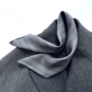 Charcoal grey silk neckerchief with light blue geometric pattern displayed on grey coat, showing luxurious sheen and hand-rolled edges