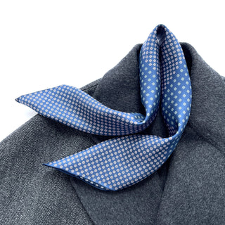 Blue silk neckerchief with white four-leaf clover pattern draped elegantly over gray wool fabric, showing hand-rolled edges and lustrous finish