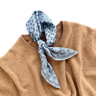 Sky blue silk neckerchief with navy paisley pattern elegantly tied around the collar of a camel-colored sweater, showcasing its hand-rolled edges.