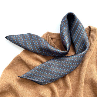 Charcoal grey silk neckerchief with light blue geometric pattern displayed on camel-colored sweater, showing luxurious texture and hand-rolled edges