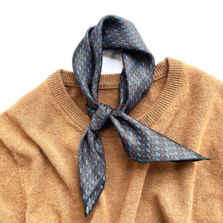 Charcoal grey silk neckerchief with light blue geometric pattern tied stylishly around the collar of a camel-colored sweater
