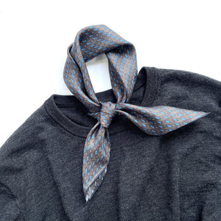 Charcoal grey silk neckerchief with light blue geometric pattern tied around dark sweater collar, showcasing luxurious sheen and texture