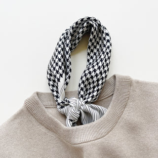 Silk scarf with black and white houndstooth pattern tied fashionably around beige sweater neckline, featuring contrast striped edge detail