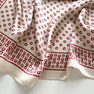 Beige silk scarf with red horseshoe pattern throughout and decorative border, showing luxurious fabric texture and elegant draping