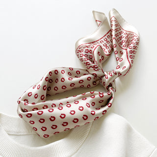 Beige silk scarf with red horseshoe pattern tied in a casual knot, displayed on white fabric showing its lustrous satin finish