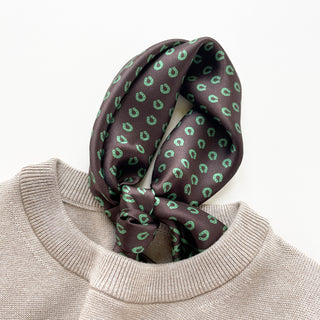 Brown silk scarf with green horseshoe pattern tied elegantly around beige sweater neckline, showcasing the shiny satin texture and classic print design