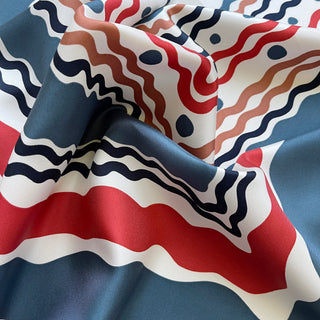 Luxury silk twill scarf with flowing wave pattern in navy, red, and earthy tones, showcasing hand-rolled edges and rich texture on a softly draped fabric.