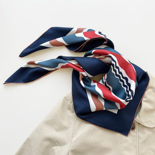 Square silk scarf with navy border and wave pattern in navy, red, blue, and terracotta colors, elegantly draped on beige fabric showing hand-rolled edges