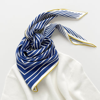 Triangular silk scarf with navy blue and white stripes, hand-rolled yellow edge, elegantly tied on white fabric, showing premium texture