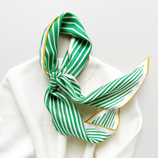 Triangle silk scarf with green and white stripes and yellow trim, elegantly tied around a white sweater neck, showing hand-rolled edges