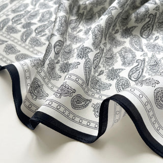 Luxurious black and white silk bandana featuring intricate paisley pattern with elegant black border trim, showing silky texture and flowing fabric