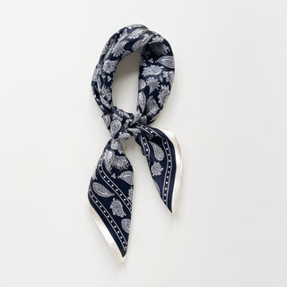 Navy blue silk bandana scarf with white paisley pattern elegantly tied in a knot, featuring decorative border and silky sheen on white background.