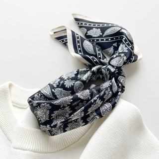 Navy blue silk bandana scarf with white paisley pattern, elegantly tied and displayed on a cream sweater collar, showing luxurious shine and texture