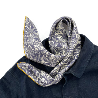 Ink blue silk scarf with gothic rose pattern and white illustrations, featuring mustard yellow hand-rolled edges, draped elegantly around dark denim collar