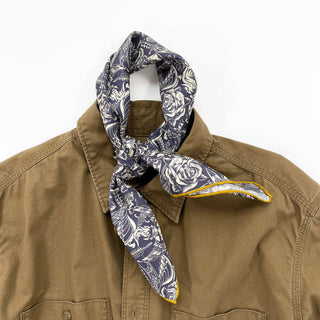 Ink blue silk scarf with white gothic rose pattern and yellow trim, tied around the collar of a brown shirt, showcasing elegant styling