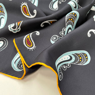 Black silk scarf with colorful paisley pattern featuring mint green, yellow, and red accents, showcasing luxurious fabric with hand-rolled yellow hem.