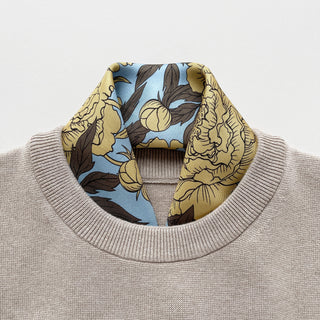Luxury silk scarf featuring light yellow peonies print on light blue background elegantly tucked  in a grey sweater on a light grey background