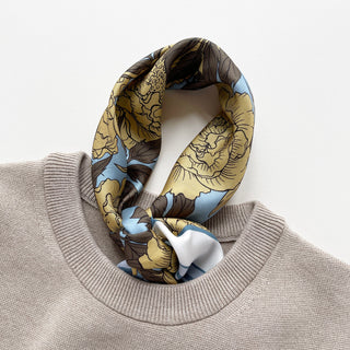 Luxury silk scarf featuring light yellow peonies print on light blue background elegantly tied in a grey sweater on a light grey background