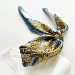 women’s silk neck scarf in light yellow peony print with dusty blue border, styled in a bow on cream white sweater.