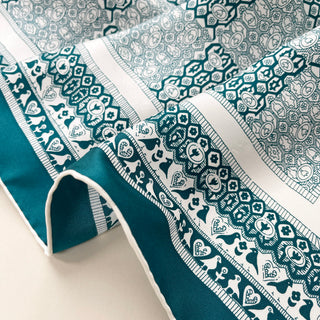 Teal silk scarf with intricate white pattern featuring birds, hearts, and geometric designs, showcasing luxurious hand-rolled edges and silky twill texture.