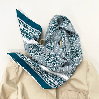 Teal Heritage Grace silk scarf with intricate white paisley patterns draped elegantly over a beige coat, showcasing its luxurious texture and hand-rolled edges