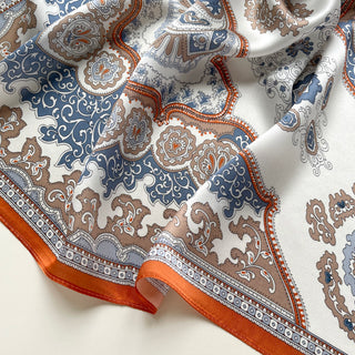 Silk scarf with intricate baroque pattern in blue, orange and beige tones, featuring ornate paisley designs and decorative orange border on luxurious fabric.
