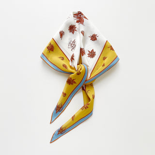 Diamond-shaped silk scarf in yellow and ivory with floral print, tied in a loose knot on a white background, showcasing blue and rust border accents.