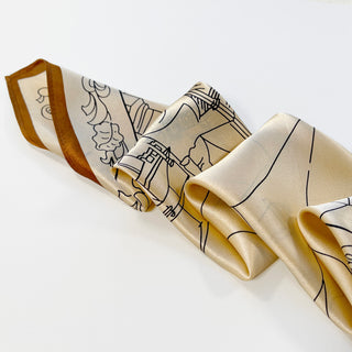 beige silk scarf with black line art cityscape design and caramel-colored border, displaying a soft sheen and elegant drape