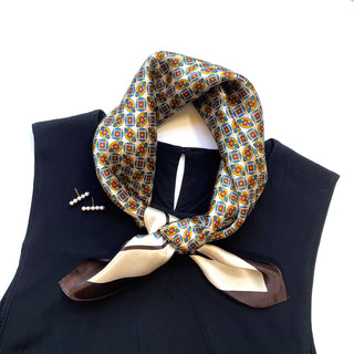 Black Heritage silk scarf with colorful geometric pattern featuring blue, red, and yellow designs, tied elegantly around a black garment with pearl earrings displayed