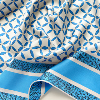 Close-up of women’s silk scarf with blue petal pattern on a light grey base, hand-rolled edges, and smooth silk twill texture.