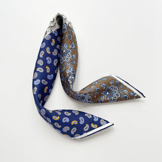 Men's silk scarf featuring floral and paisley patterns in blue and olive brown with navy blue hand-rolled edges on a light gray background.