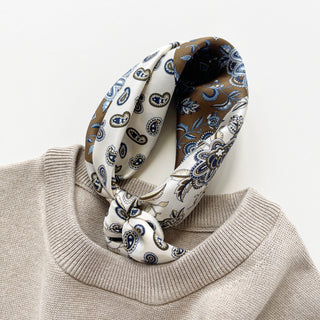 men's silk neckerchief with paisley and floral patterns in cream and olive brown knotted elegantly around the neckline of a beige sweater on a white background