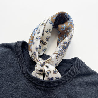 men's silk neckerchief with paisley and floral patterns in cream and olive brown knotted elegantly around the neckline of a dark grey sweater on a white background
