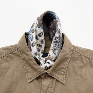 men's silk neckerchief with paisley and floral patterns in cream and olive brown knotted elegantly around the neckline of a brown shirt on a white background