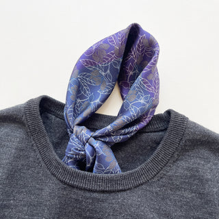 Men’s silk neckerchief scarf featuring blue and purple floral print with hand-rolled edges, tied around the neck line of a grey sweater.