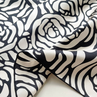 Close up of charcoal black and cream white silk scarf  with a rose print design, showing luxurious fabric texture and hand-rolled edge on a light grey background