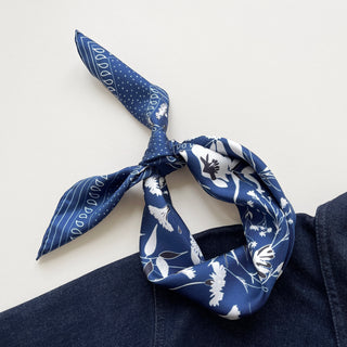 Blue Kyoto Blooms silk scarf tied as a bandana, featuring white floral patterns, polka dots, and stripes on luxurious silk twill fabric against denim