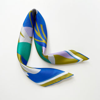 Luxurious abstract lotus silk scarf in vibrant blue, teal, lilac, and mustard yellow patterns with hand-rolled edges, elegantly draped on a light grey background.