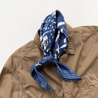 Blue Kyoto Blooms silk scarf with white floral pattern tied as a bandana on tan button-up shirt collar, showcasing hand-rolled edges