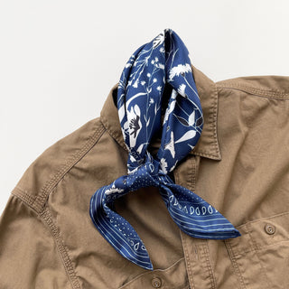 Blue Kyoto Bloom silk neckerchief with white botanical floral pattern, elegantly styled on a tan button-up shirt collar.