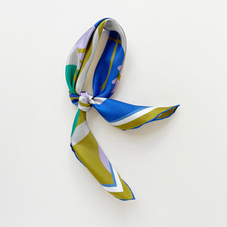 Luxurious abstract lotus silk scarf in vibrant blue, teal, lilac, and mustard yellow patterns with hand-rolled edges, elegantly tied on a light grey background.